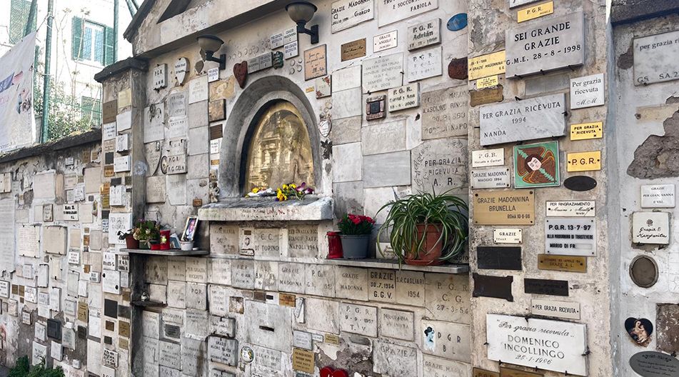 Eine mit zahlreichen Danktafeln bedeckte Wand eines kleinen Schreins oder Altars im Freien. In der Mitte befindet sich eine vergoldete Marienfigur hinter Glas, umgeben von Blumen, Kerzen und Pflanzen. Die Tafeln tragen italienische Inschriften mit Danksagungen für erhörte Gebete („Per grazia ricevuta“).