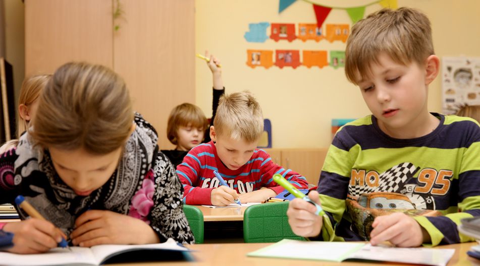Mehrere Grundschulkinder sitzen in einem bunt dekorierten Klassenzimmer an ihren Tischen und schreiben konzentriert in ihre Hefte. Ein Kind im Hintergrund hebt die Hand.